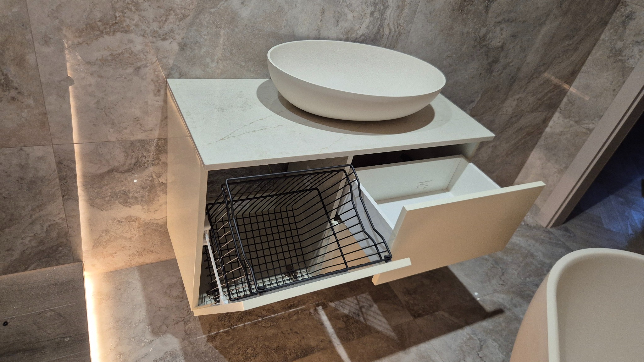 Nowoczesna szafka łazienkowa z blatem imitującym marmur i owalną umywalką nablatową. Otwarta szuflada z metalowym koszem i druga z białym wnętrzem. Minimalistyczny design.