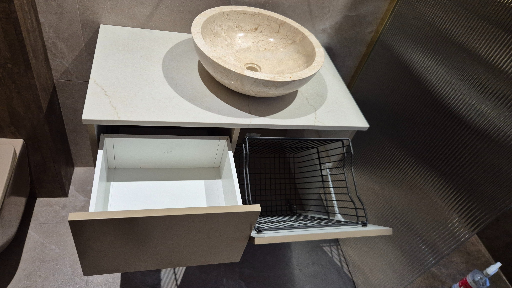 Beżowa umywalka nablatowa na blacie z otwartą szufladą i koszem cargo. Nowoczesna łazienka z dekoracyjną ścianą. Ujęcie z góry.