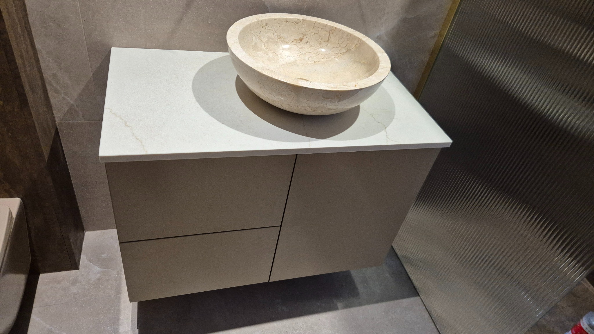 Beżowa szafka łazienkowa z blatem z jasnego kamienia i umywalką w kształcie misy z marmuru, na tle ściany z dekoracyjną fakturą.