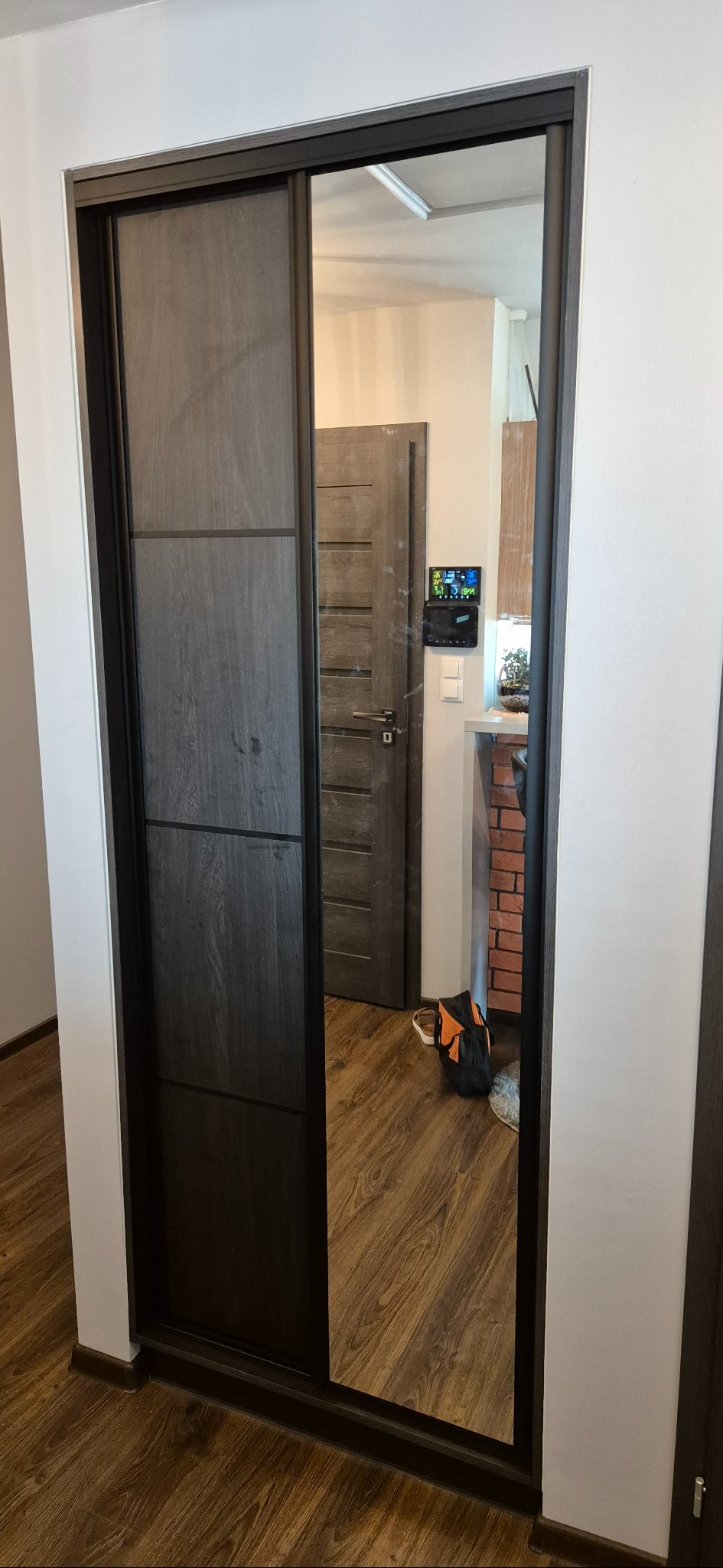 Szafa wnękowa z przesuwnymi drzwiami z lustrem, w odcieniach szarości, z widoczną strukturą drewna. W lustrze odbija się wnętrze mieszkania.