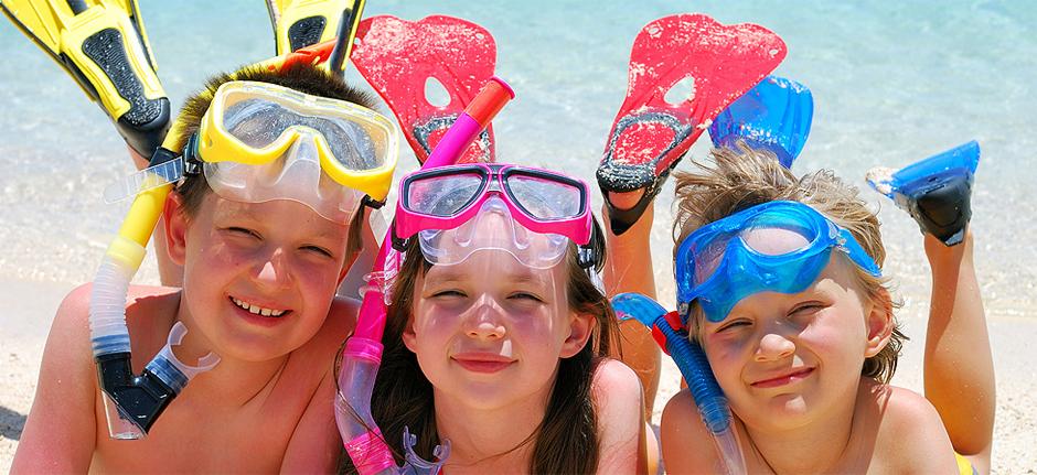 Troje uśmiechniętych dzieci w maskach do nurkowania i płetwach leży na piaszczystej plaży, z widokiem na morze w tle.