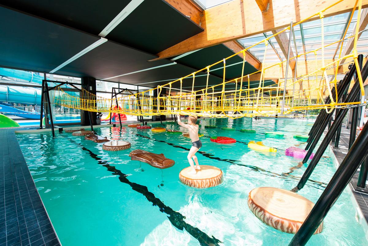 Chłopiec w kąpielówkach skacze po pływających platformach imitujących pnie drzew w basenie z atrakcjami wodnymi, nad nim żółta siatkowa konstrukcja linowa, w tle zjeżdżalnie wodne.