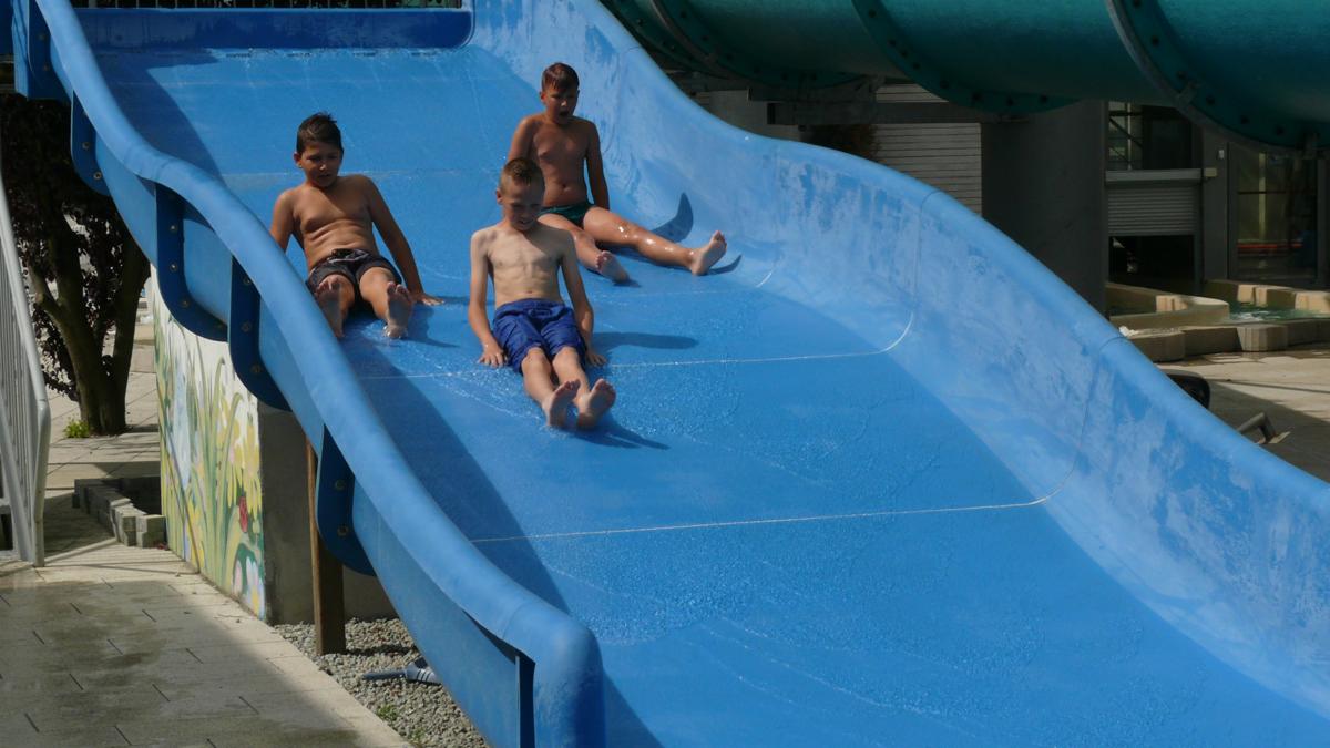Trójka dzieci zjeżdża na niebieskiej zjeżdżalni wodnej w aquaparku, widoczne płytki basenowe i fragment rury zjeżdżalni w tle.