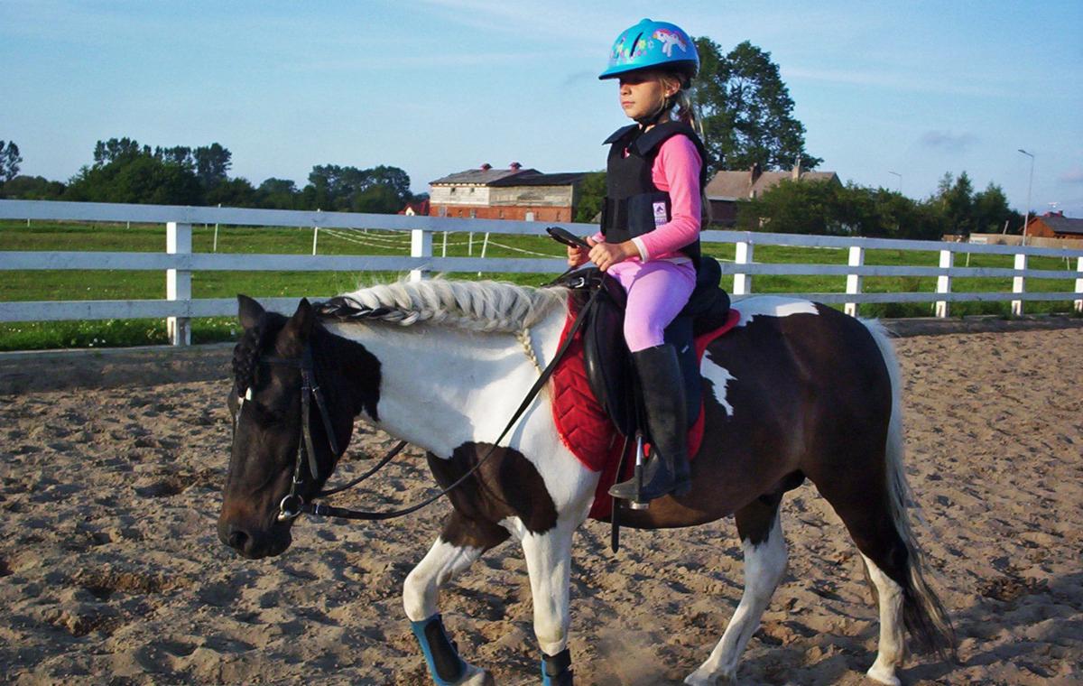 Dziewczynka w niebieskim kasku z jednorożcami i kamizelce ochronnej jeździ na łaciatym koniu z zaplecioną grzywą na piaszczystym padoku.