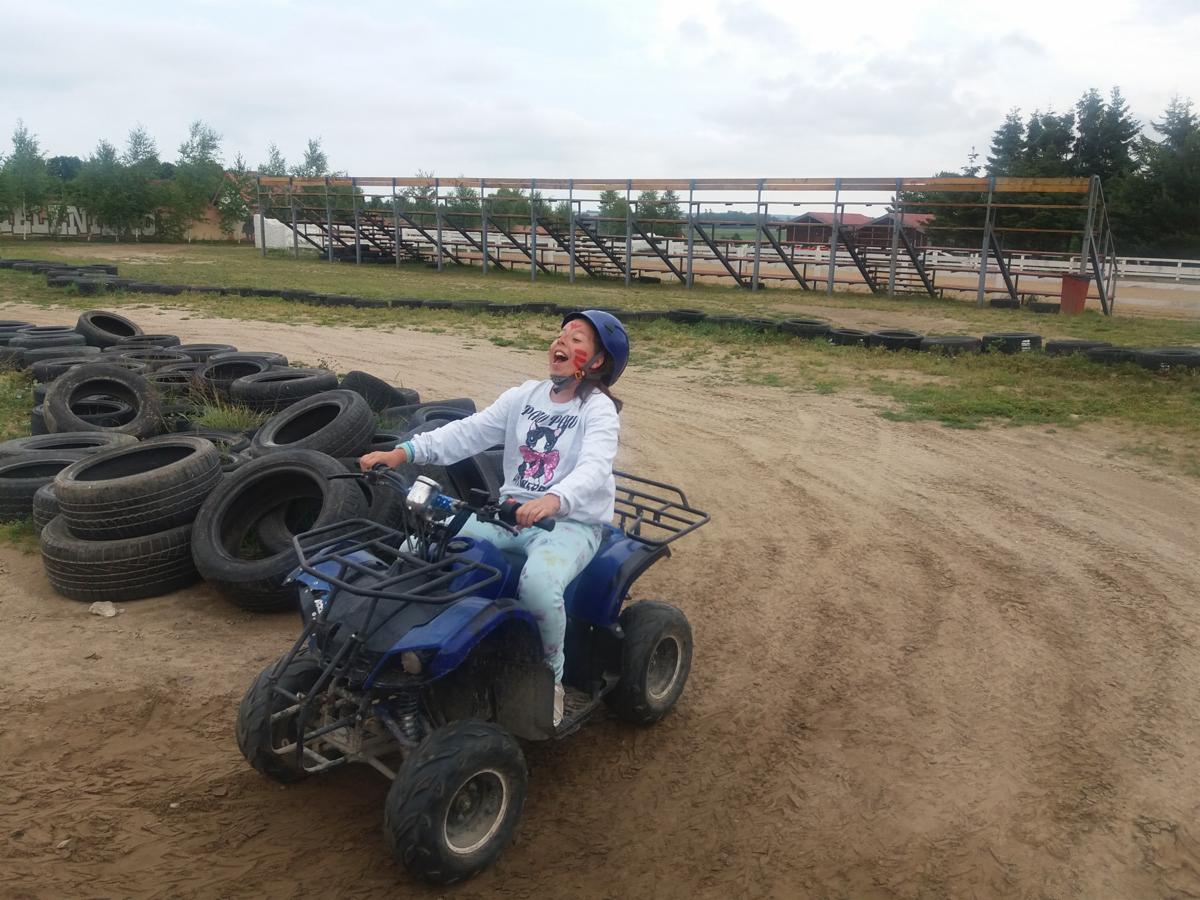 Dziewczynka w kasku i z pomalowaną twarzą, z szerokim uśmiechem, siedzi na niebieskim quadzie na piaszczystym torze z oponami i trybunami w tle.