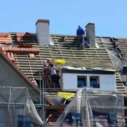 Ekipa dekarzy na rusztowaniu podczas wymiany poszycia dachu, widoczne fragmenty starej i nowej konstrukcji, kominy oraz zabezpieczenia rusztowania.