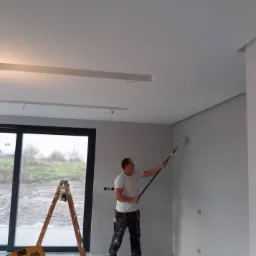 Mężczyzna maluje szarą ścianę wałkiem na teleskopowym drążku w jasnym pomieszczeniu z oknem, widoczna drabina, grzejnik i nieotynkowany sufit z listwami.