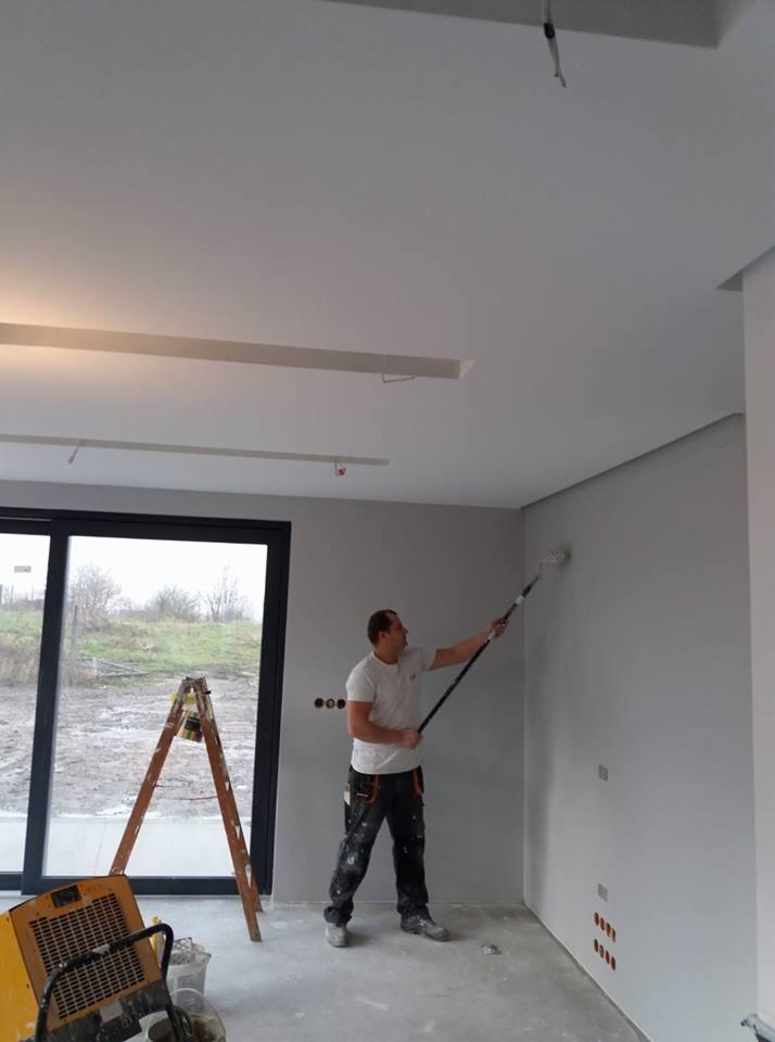 Mężczyzna maluje szarą ścianę wałkiem na teleskopowym drążku w jasnym pomieszczeniu z oknem, widoczna drabina, grzejnik i nieotynkowany sufit z listwami.