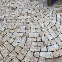 Fragment układanej nawierzchni z kostki granitowej w kształcie łuku, widoczne czerwone sznurki wyznaczające linię układania oraz koło taczki w rogu.