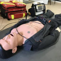 Manekin ułożony na macie, ubrany w robocze spodnie i rozpiętą kurtkę, podłączony do defibrylatora, obok torba ratownicza i urządzenie medyczne.
