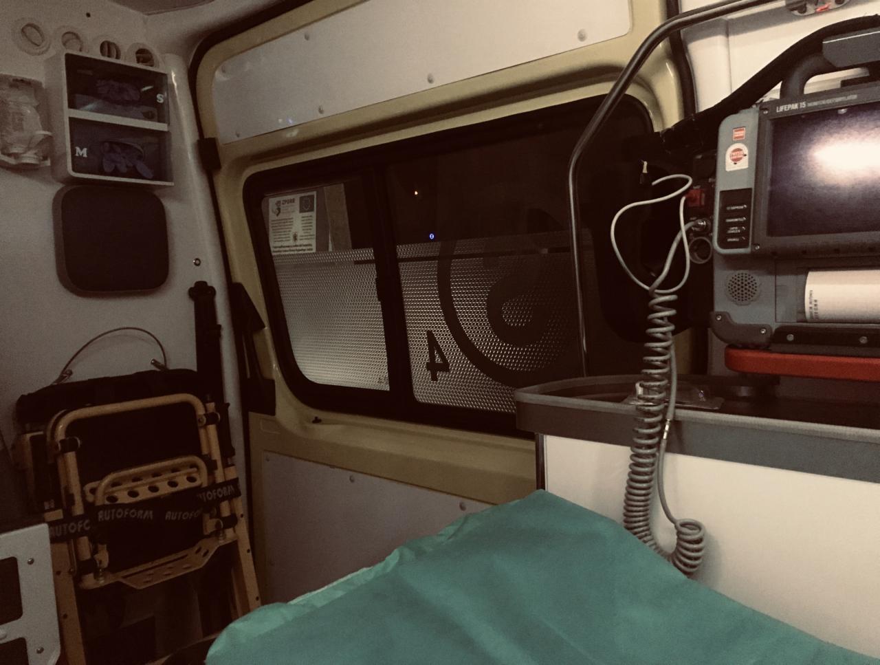 Wnętrze ambulansu: składane krzesło transportowe, defibrylator LifePak 15, szafka z wyposażeniem medycznym i okno z kratką zabezpieczającą.