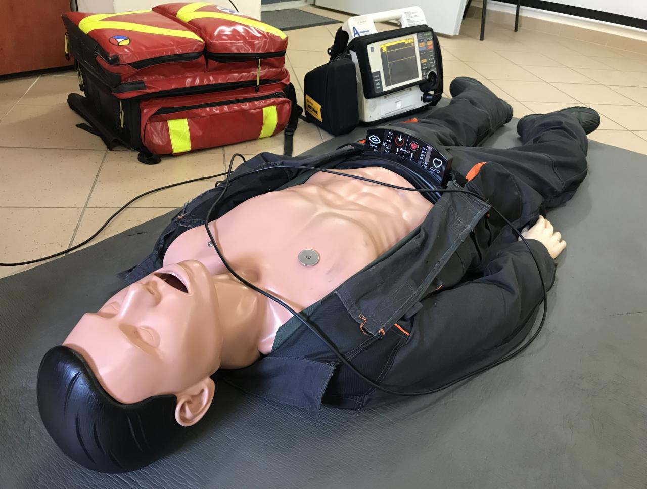Manekin ułożony na macie, ubrany w robocze spodnie i rozpiętą kurtkę, podłączony do defibrylatora, obok torba ratownicza i urządzenie medyczne.
