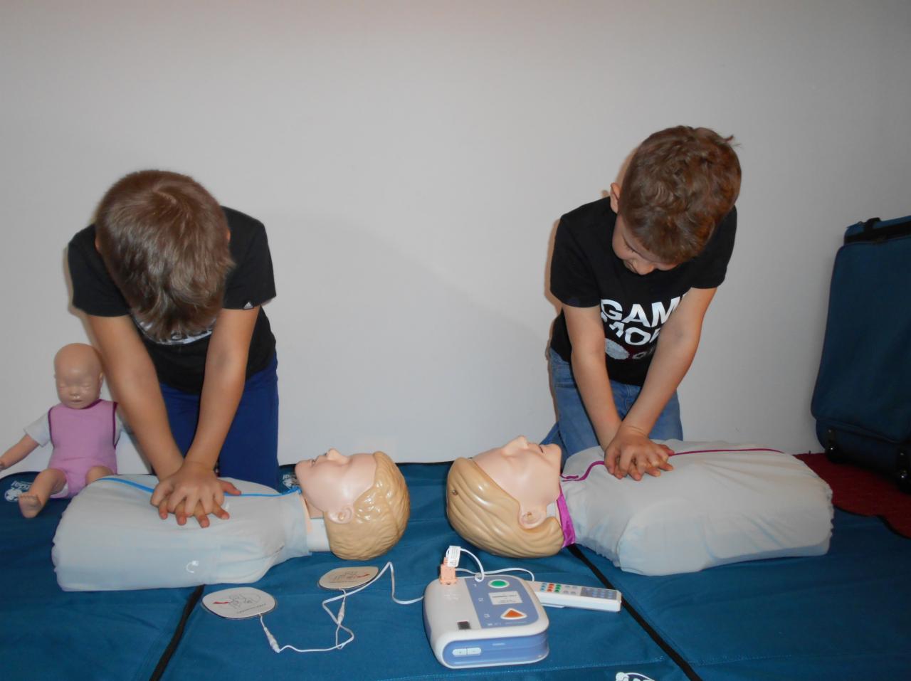 Dwaj chłopcy w czarnych koszulkach ćwiczą resuscytację krążeniowo-oddechową na manekinach z blond włosami, obok defibrylator i lalka w różowym ubranku.