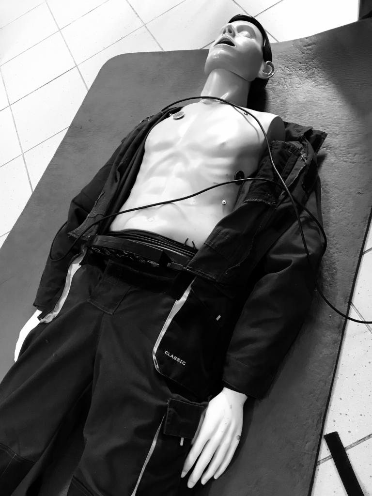 Manekin szkoleniowy leżący na plecach, ubrany w kombinezon roboczy z napisem 'CLASSIC', z podłączonymi elektrodami EKG, na kafelkowanej podłodze.