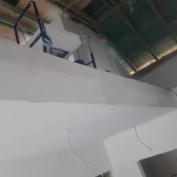 Wnętrze w trakcie prac wykończeniowych, widoczny sprzęt do natryskowego nakładania gładzi Texspray T-Max na tle surowego, drewnianego stropu i świeżo pomalowanych ścian.