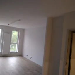 Pusty, jasny pok&oacute;j po remoncie z drewnianą podłogą, białymi ścianami, dwoma oknami z widokiem na zieleń i grzejnikiem pod oknem, widoczne gniazdka elektryczne przy podłodze.