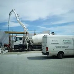 Betonomieszarka z pompą podająca beton na budowę domu, widoczny fragment szalunku i zaparkowany biały bus firmy budowlanej.