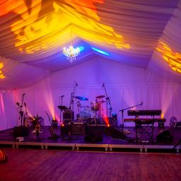 Go Art - Scena muzyczna z instrumentami (perkusja, gitary, saksofon, keyboard) i nagłośnieniem wewnątrz białego namiotu oświetlonego kolorowymi światłami, z żyrandolem na suficie.