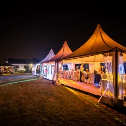 Oświetlony namiot bankietowy z drewnianą podłogą i białymi krzesłami ustawionymi do uroczystej kolacji, widoczny z zewnątrz w nocnym ogrodzie, z dodatkowym oświetlonym pawilonem w tle.