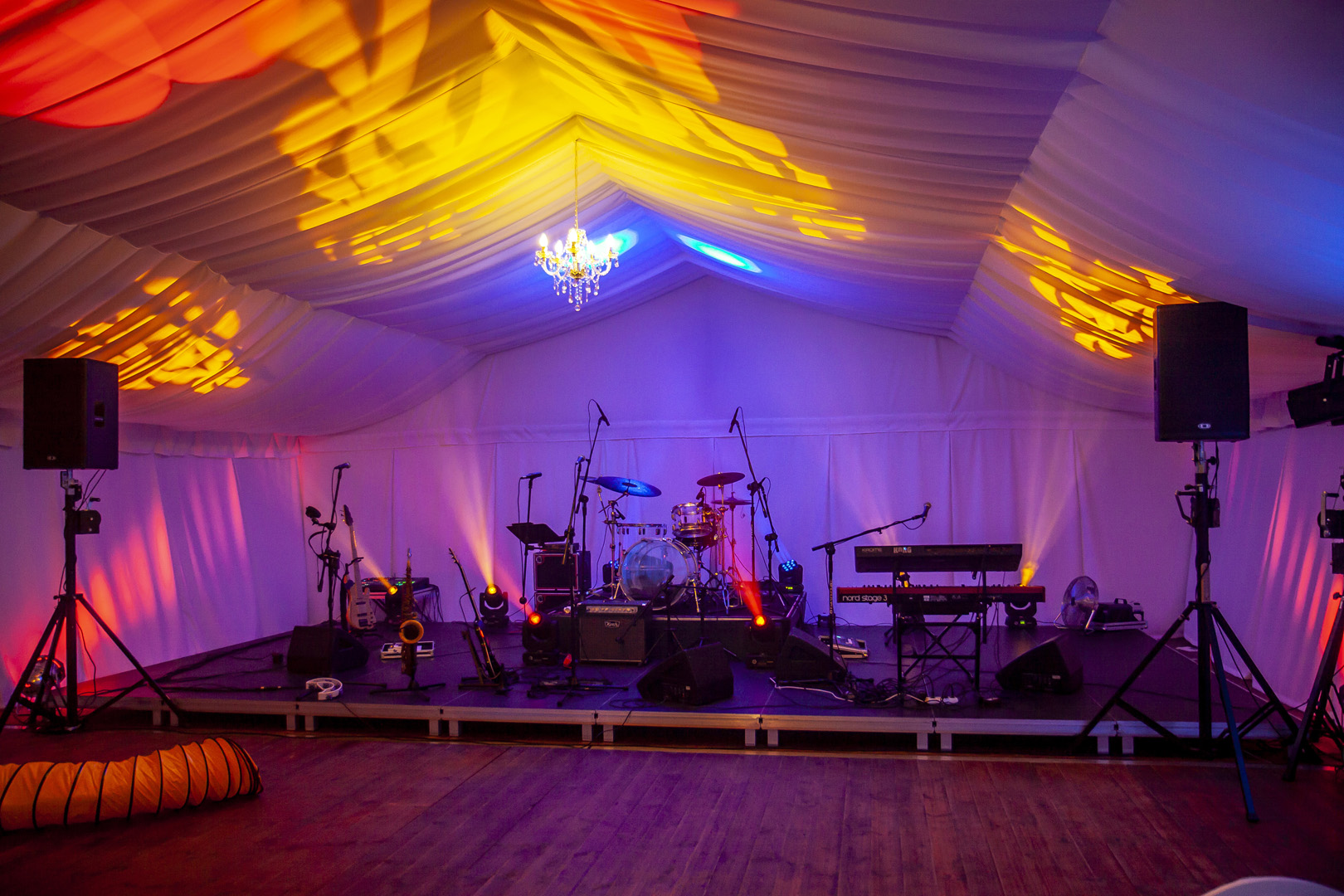 Scena muzyczna z instrumentami (perkusja, gitary, saksofon, keyboard) i nagłośnieniem wewnątrz białego namiotu oświetlonego kolorowymi światłami, z żyrandolem na suficie.