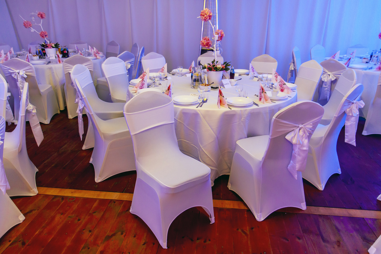 Uroczysty stół z białym obrusem, nakryciami i dekoracjami kwiatowymi, otoczony krzesłami w białych pokrowcach z różowymi akcentami, ustawiony wewnątrz namiotu z drewnianą podłogą.