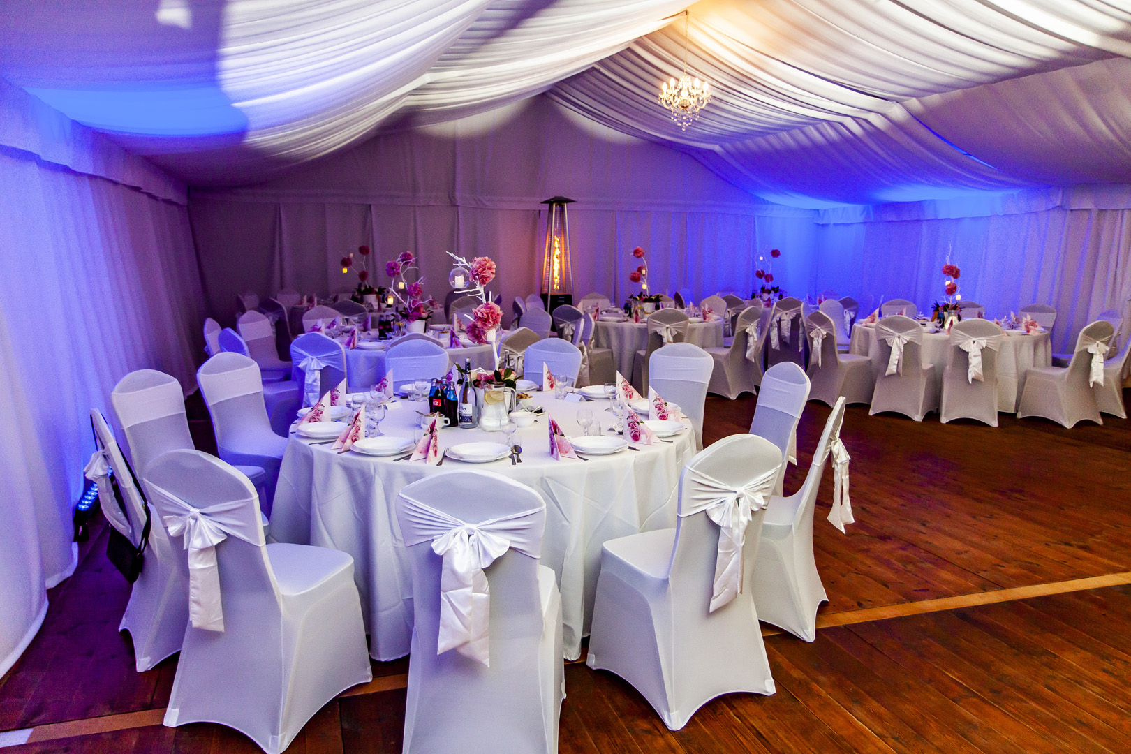 Uroczyste wnętrze białego namiotu bankietowego z okrągłymi stołami, krzesłami z białymi pokrowcami i kokardami, dekoracjami kwiatowymi, oświetlone niebieskim światłem i wiszącym kryształowym...