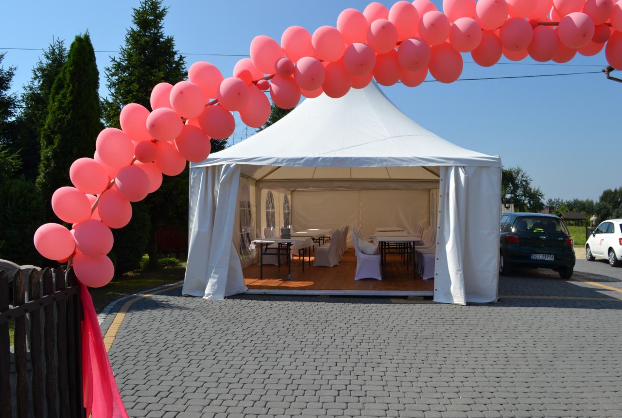 Biały namiot imprezowy udekorowany łukiem z różowych balonów, z widocznymi stolikami i krzesłami wewnątrz, ustawiony na brukowanym podjeździe w słoneczny dzień.