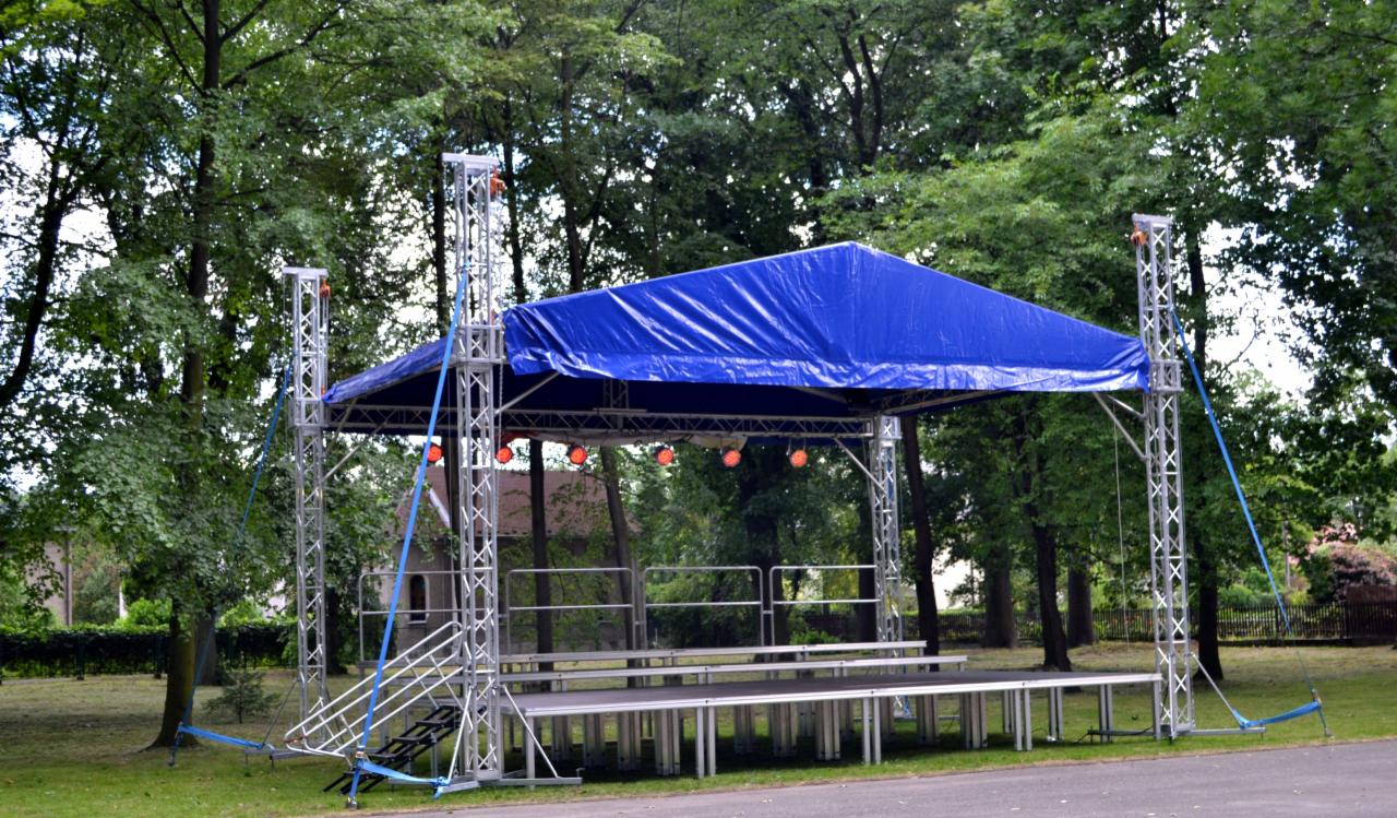 Scena plenerowa z niebieskim zadaszeniem, oświetleniem punktowym i metalową konstrukcją, otoczona zielenią drzew.