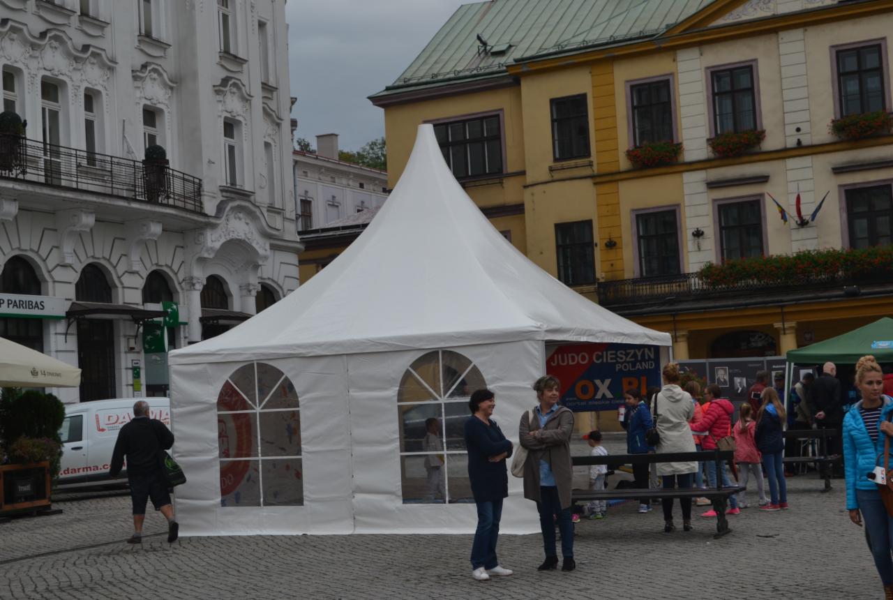 Biały namiot z oknami w kształcie łuku, ustawiony na brukowanym placu w otoczeniu budynków o zróżnicowanej architekturze, z widocznymi osobami w tle.