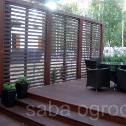 Zadaszony taras z drewnianą podłogą i ażurową ścianką, zestaw mebli ogrodowych z ciemnego rattanu i szklanym blatem, donice z zielenią.