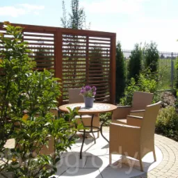 Aranżacja przestrzeni ogrodowej z drewnianą przesłoną, okrągłym stołem z kwiatami i rattanowymi krzesłami, na tle zieleni i kostki brukowej ułożonej w łuk.