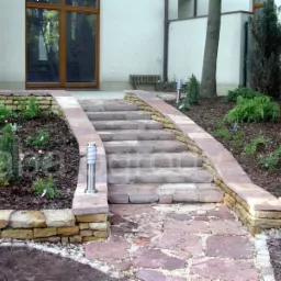 Schody z kamienia prowadzące do domu, otoczone rabatami z roślinami i korą, ścieżka z nieregularnych płyt kamiennych.