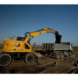 Żółta koparka kołowa CAT M315 załadowuje urobek do skrzyni ciężarówki na placu budowy w słoneczny dzień, w tle widoczne pole i słupy energetyczne.