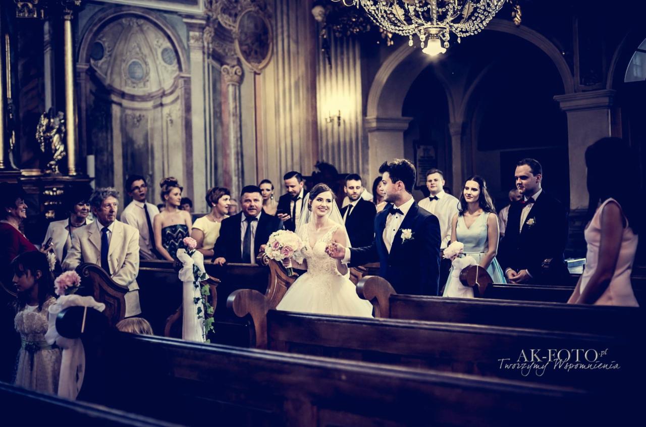 Para młoda w kościele, panna młoda w białej sukni z bukietem, pan młody w ciemnym garniturze, goście weselni siedzą w ławkach, żyrandol nad głową.