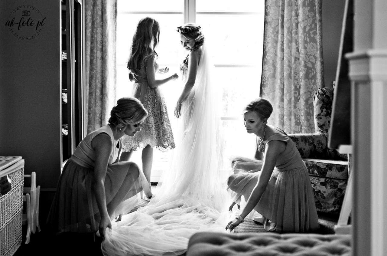 Czarno-białe zdjęcie panny młodej w sukni ślubnej z welonem, otoczonej przez trzy druhny przygotowujące się do ceremonii przy jasnym oknie.