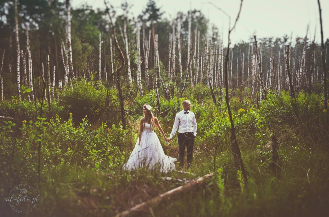 Para młoda trzymająca się za ręce podczas sesji w lesie z brzozami, panna młoda w białej sukni z kwiatowym wiankiem, pan młody w białej koszuli i czarnej muszce.