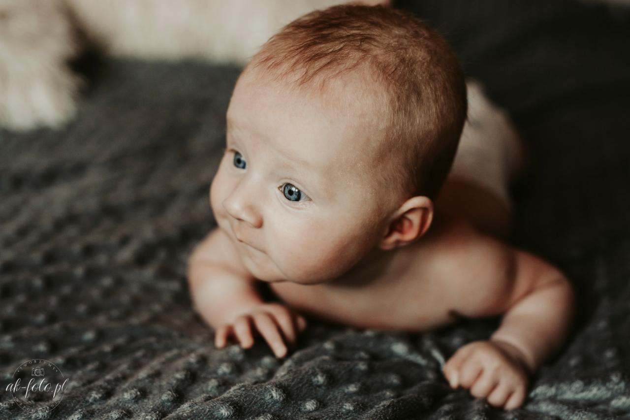Noworodek z jasną cerą i niebieskimi oczami leży na brzuchu na szarym, teksturowanym kocyku, spoglądając w lewo.