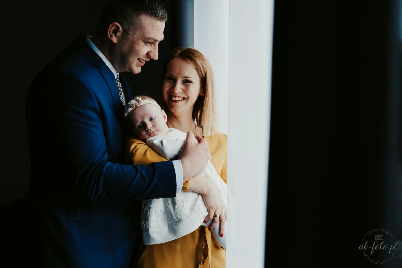 Uśmiechnięta rodzina z niemowlęciem w białej sukience i opasce z kwiatami, ojciec w granatowym garniturze, matka w musztardowej sukience, stoją przy oknie.