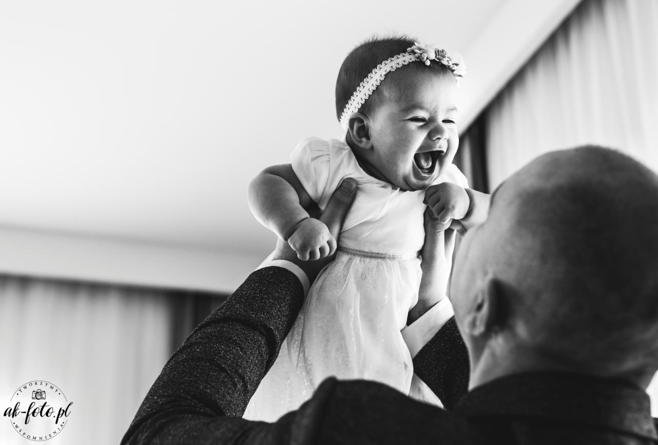 Czarno-białe zdjęcie niemowlęcia w białej sukience i opasce na głowie, trzymanego w górze przez mężczyznę w garniturze, z szerokim uśmiechem i otwartymi ustami, uchwycone z perspektywy mężczyzny.