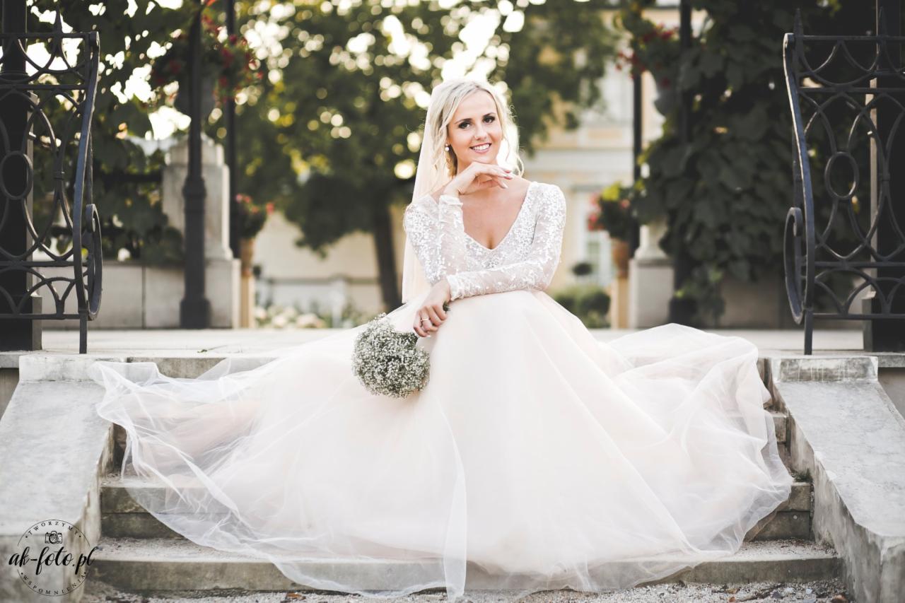 Panna młoda w sukni z długim rękawem ozdobionym cekinami, siedząca na kamiennych schodach z bukietem gipsówki, w tle park i budynek.