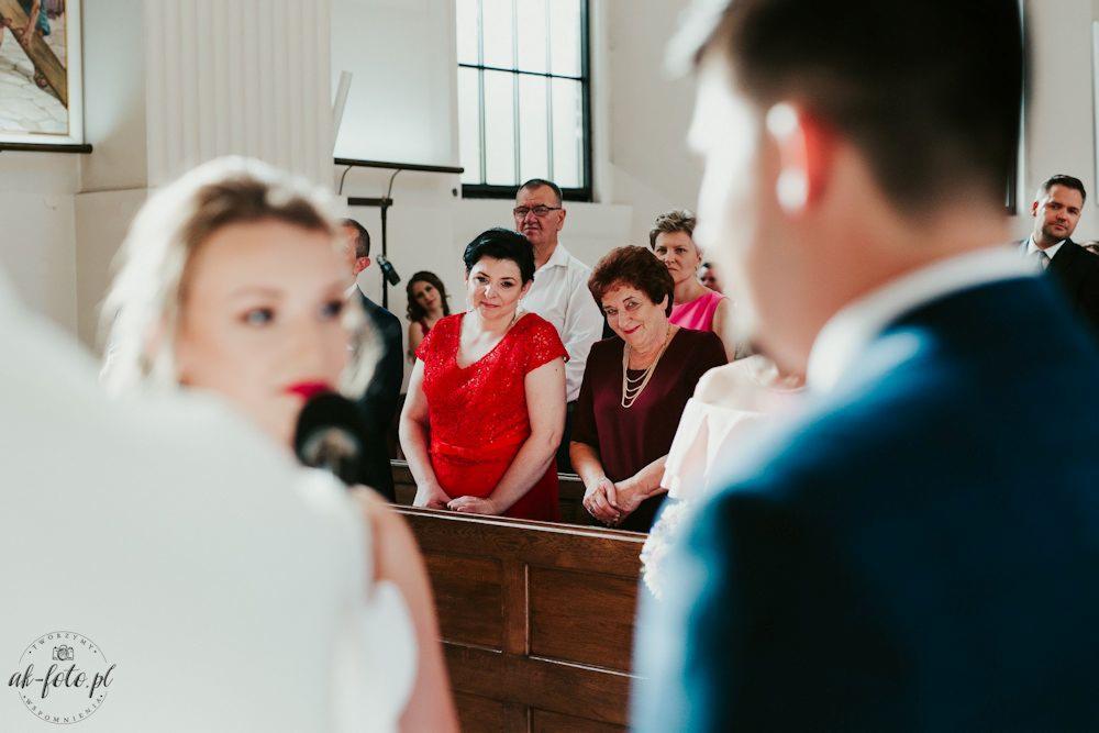 Goście weselni obserwujący parę młodą w kościele, kobiety w czerwonej i bordowej sukience z biżuterią, w tle okno i inni goście.