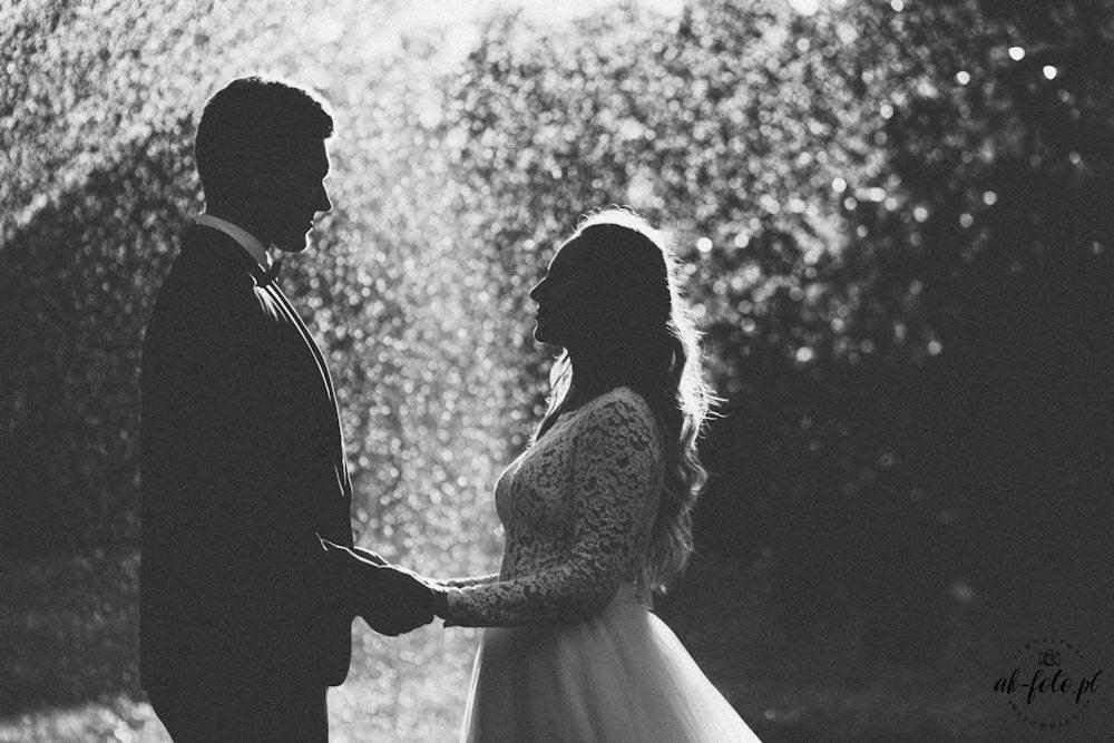 Czarno-białe zdjęcie sylwetek pary młodej trzymającej się za ręce na tle rozmytych świateł, panna młoda w koronkowej sukni z długim rękawem.