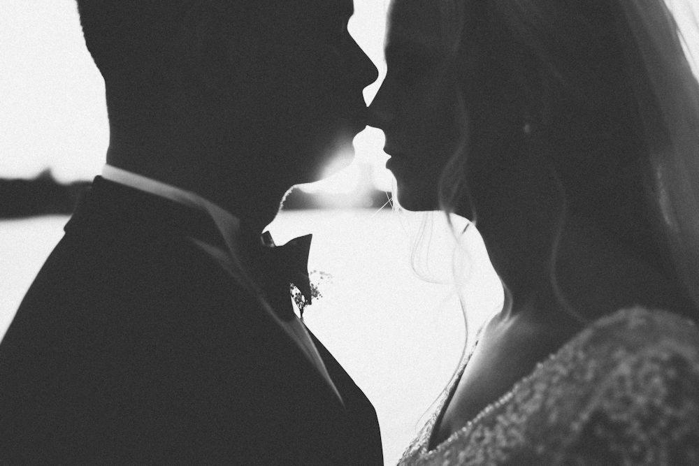 Czarno-białe zdjęcie sylwetek całującej się pary młodej na tle jasnego, rozmytego krajobrazu, z widoczną muszką pana młodego i zdobieniami sukni panny młodej.