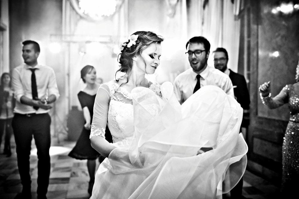 Czarno-białe zdjęcie panny młodej w koronkowej sukni z kwiatami we włosach, trzymającej falujący dół sukni podczas weselnego przyjęcia, z gośćmi w tle.