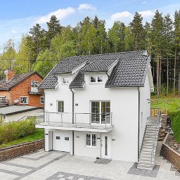 Ata Development - Nowoczesny, dwupiętrowy dom z białymi ścianami i ciemnoszarą dachówką, balkon z szklaną balustradą, garaż w bryle budynku, otoczony zielenią i innymi domami.