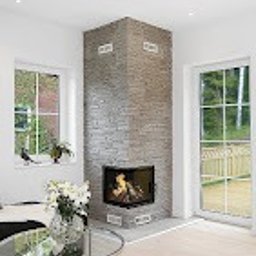 Ata Development - Wnętrze salonu z narożnym kominkiem obłożonym szarą cegłą, płomienie widoczne za szybą, jasne ściany i podłoga, okno z widokiem na zieleń, szklany stolik kawowy z bukietem kwiatów.