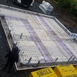 Ata Development - Przygotowanie płyty fundamentowej z instalacją ogrzewania podłogowego, widoczne rury grzewcze ułożone na warstwie izolacyjnej, zbrojenie z siatki stalowej, szalunek obwodowy i stos bloczków Ytong.