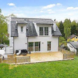 Ata Development - Nowoczesny dom z białymi ścianami, czarnym dachem z dachówki karpiówki i drewnianym tarasem z ażurową balustradą, widok z ogrodu w słoneczny dzień.