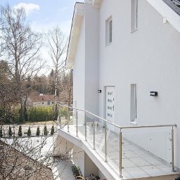 Ata Development - Biały dom z balkonem z przeszkloną balustradą, widoczna również pomarańczowa betoniarka pod balkonem i fragment dachu z szarej dachówki.