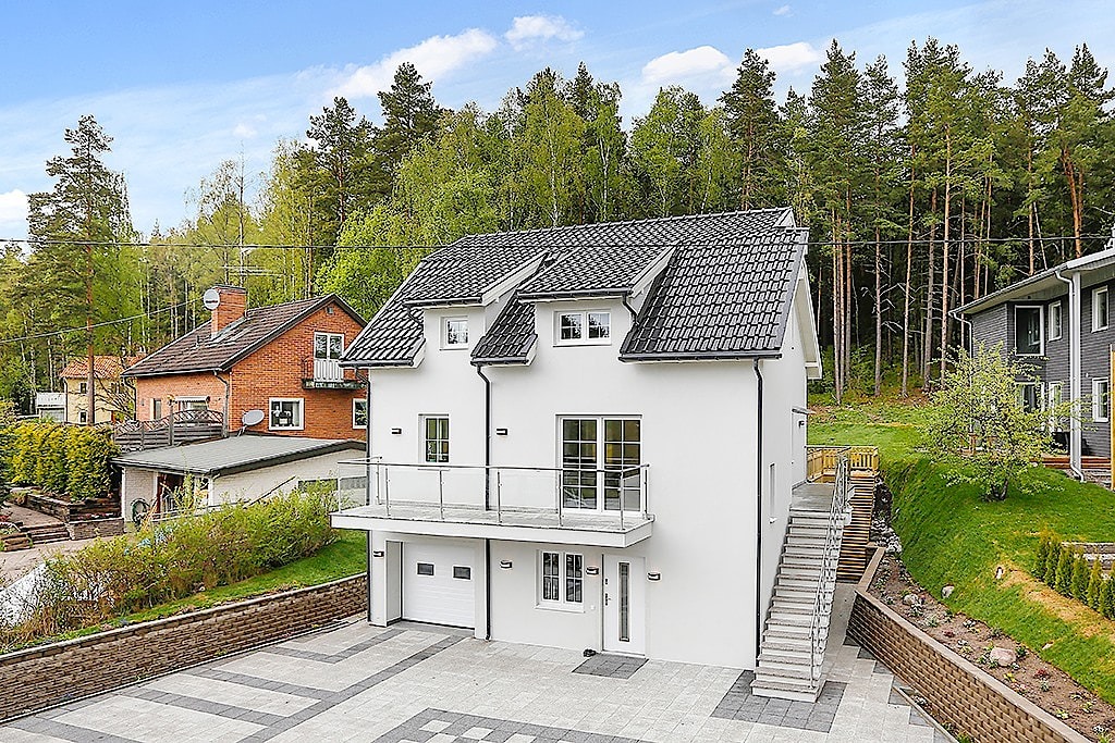 Nowoczesny, dwupiętrowy dom z białymi ścianami i ciemnoszarą dachówką, balkon z szklaną balustradą, garaż w bryle budynku, otoczony zielenią i innymi domami.